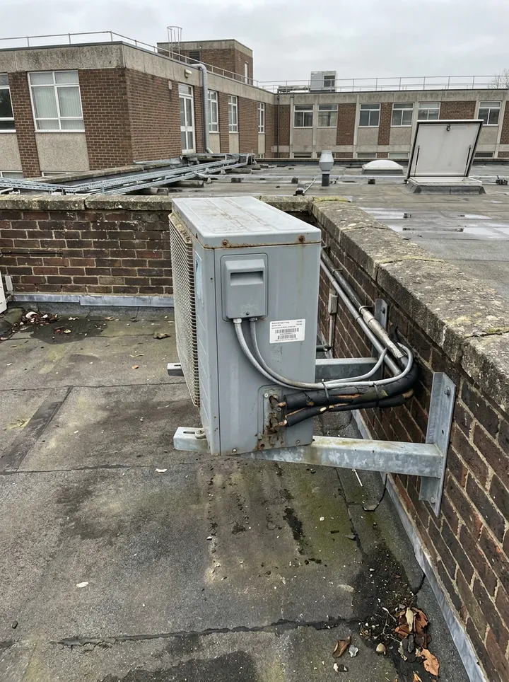 A grey air conditioning condensing unit on wall brackets beside a brick parapet on a flat roof
