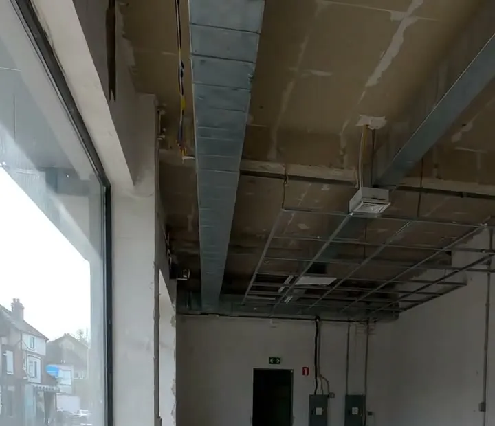 A suspended ceiling grid with most tiles out, ductwork and an air conditioning grille