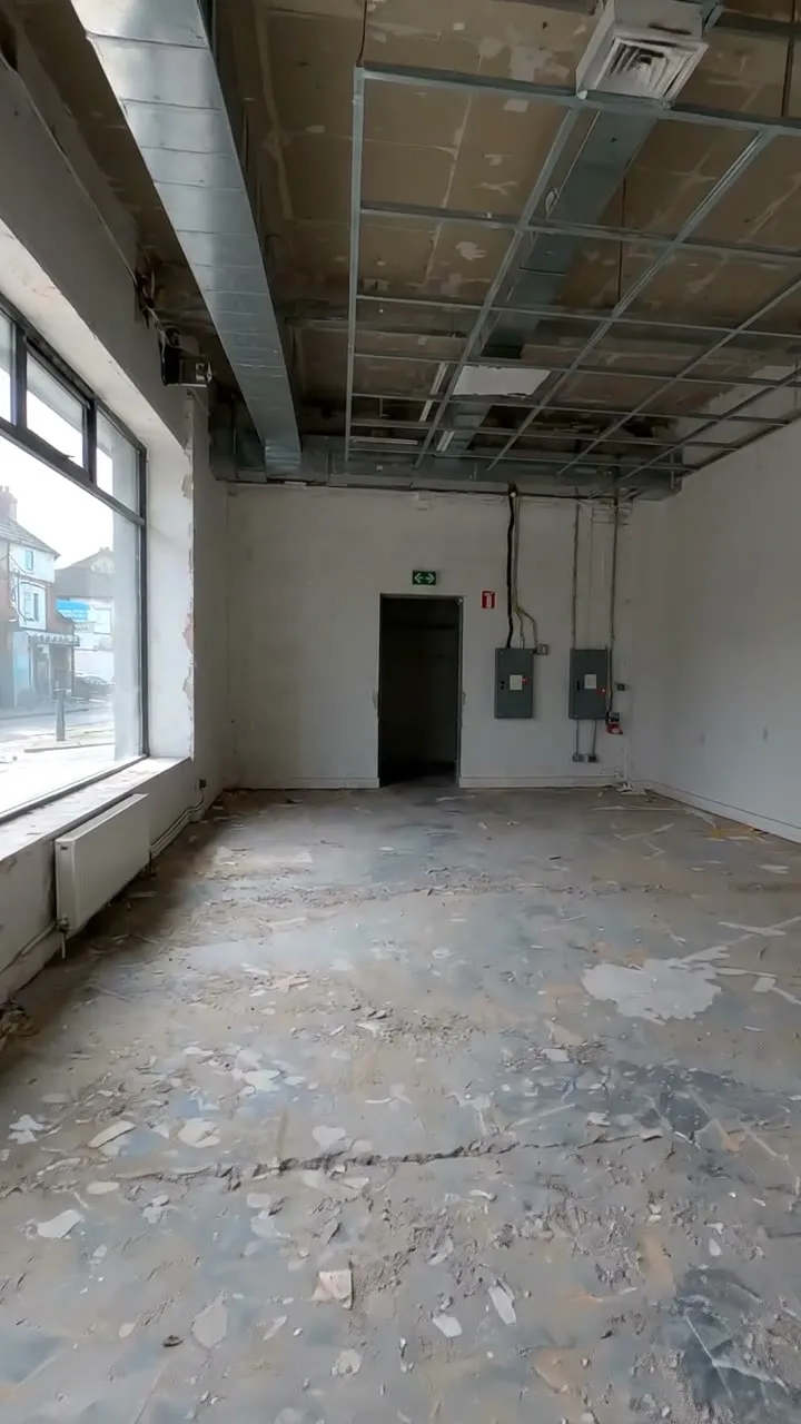 An empty former shop floor with a suspended ceiling grid, ductwork, a rear door and two distribution boards on the back wall