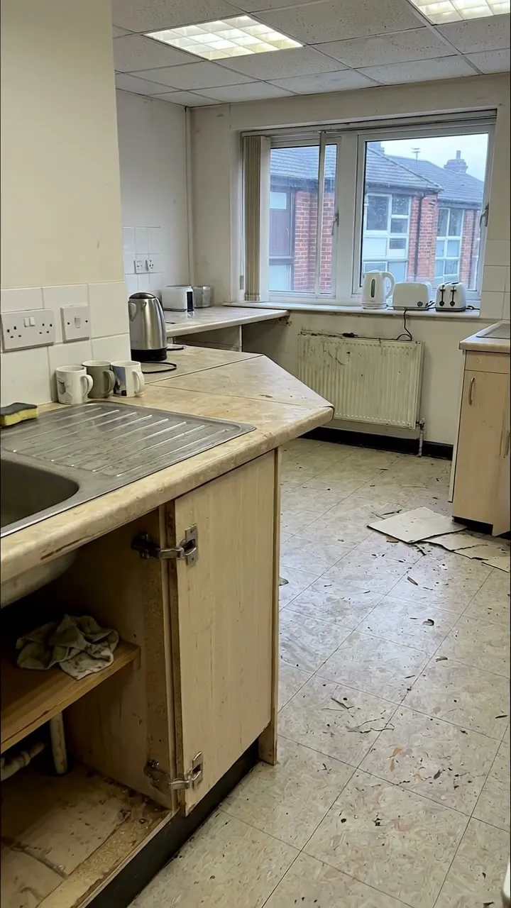 A tea point with a dirty floor, debris by the units and an open cupboard under the sink