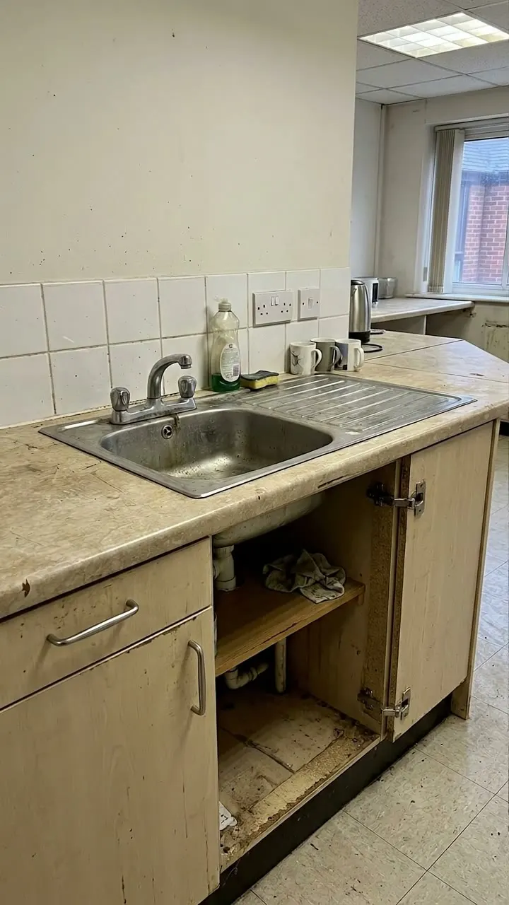 Office kitchenette with a worn worktop edge and a water-damaged sink base unit with its doors missing