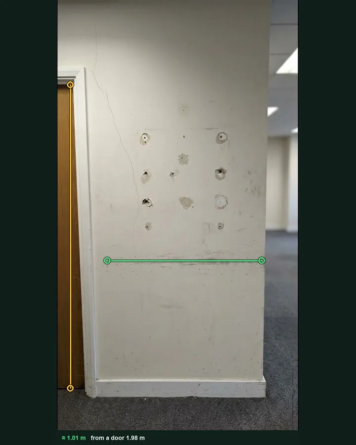 An office wall with fixing holes and a scuffed band marked with a green measuring line, against an amber line on the door beside it, giving about 1.01 m