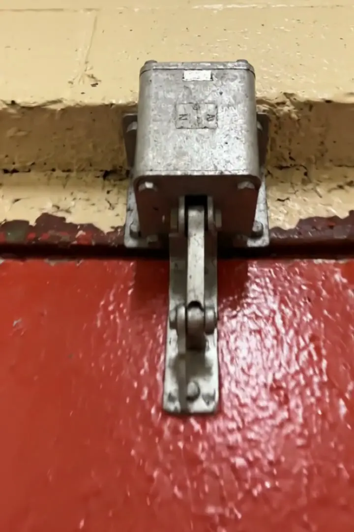 An overhead door closer arm hanging loose from its bracket above a red fire door