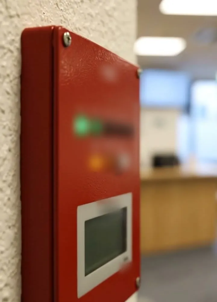 A red fire alarm panel on a textured wall, with a green indicator lit and a small display