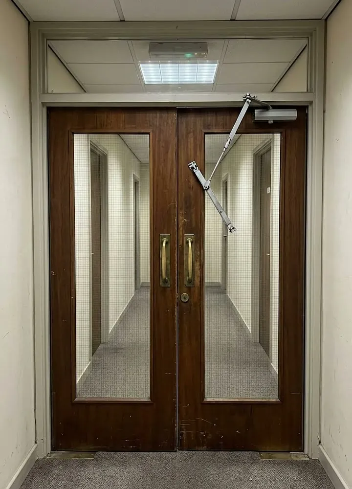 A pair of fire doors across an office corridor with an overhead door closer arm hanging loose
