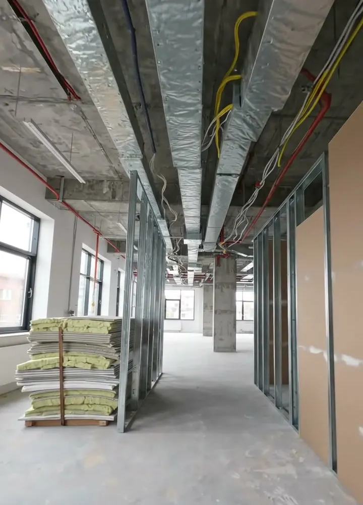 Metal stud partitions framed out along an office corridor under exposed ductwork, with plasterboard stacked on the floor