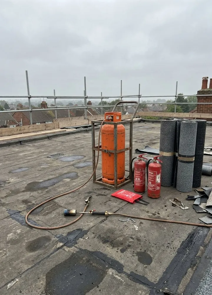 A hot works station on a flat roof: an orange propane cylinder strapped upright in a frame, two red water extinguishers, a fire blanket and rolls of torch-on felt