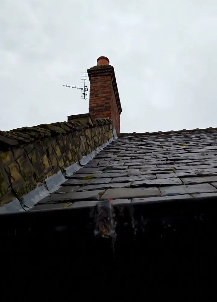 Looking up a slate roof slope towards a brick chimney stack with a TV aerial, under a grey sky