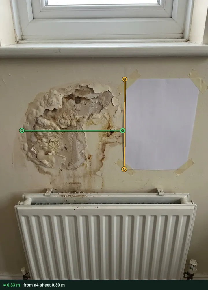 Blown plaster and tide marks on a living room wall above a radiator, measured from a photo at about 0.33 m wide using an A4 sheet