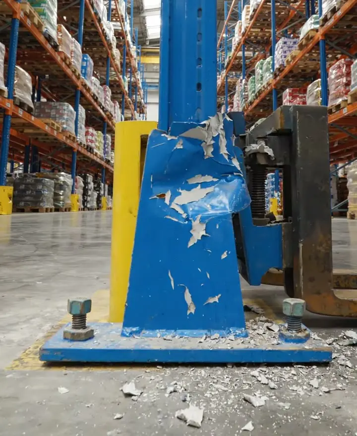 A blue pallet racking upright dented and bent just above its base plate, with loaded pallets above