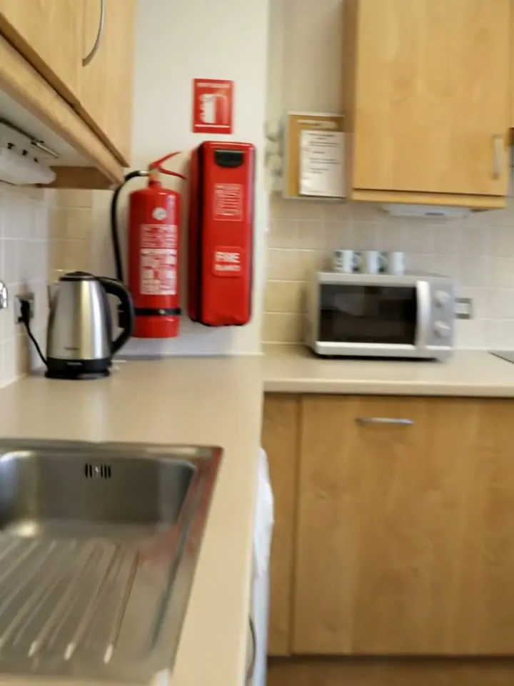 A red fire extinguisher and a fire blanket on the wall of a staff kitchen beside a kettle