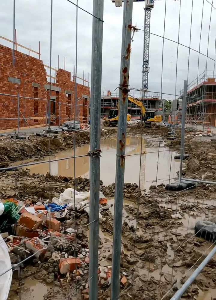 A Heras fence panel by a gate with the coupler held by tie wire, a muddy site behind