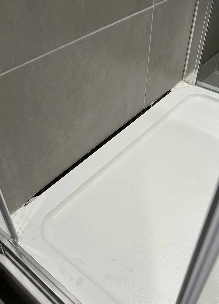 A white shower tray against grey tiles with the silicone missing along the back edge