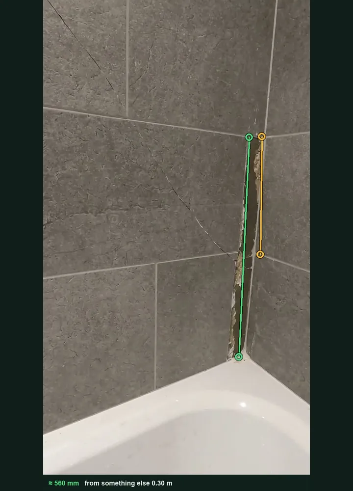 A bathroom tile corner marked up in Measure from photo: an amber line on one 300 mm tile and a green line along the open grout joint, about 560 mm