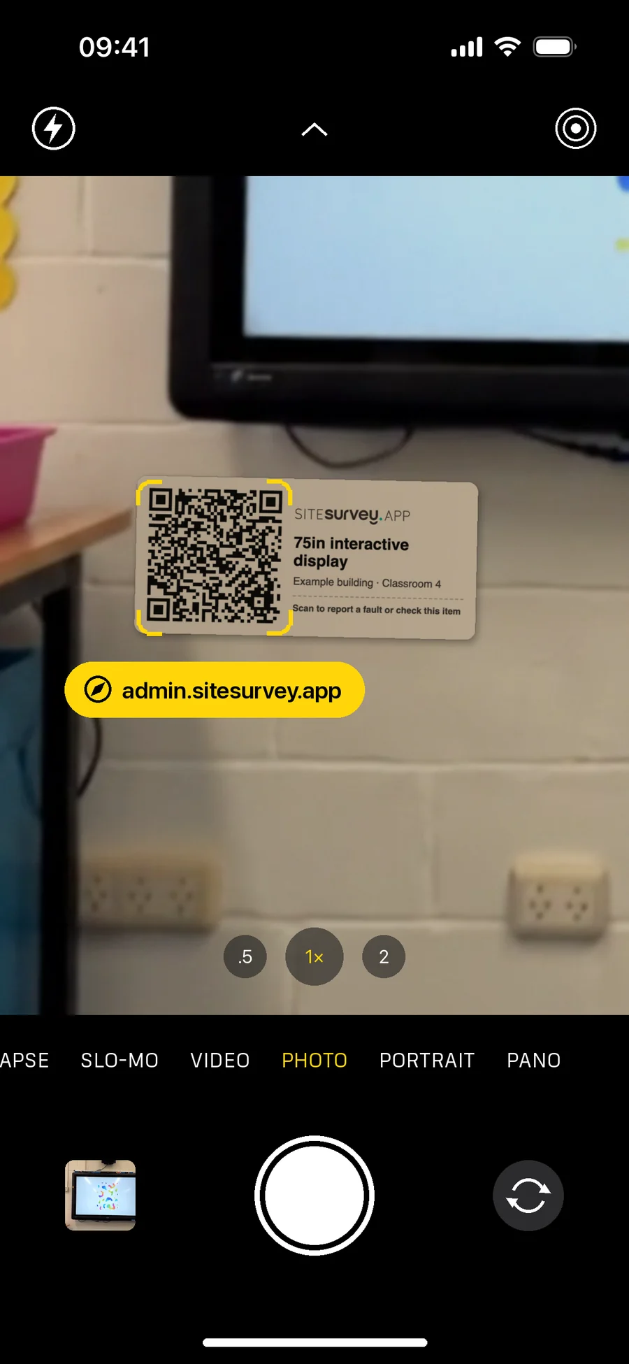 The iPhone camera pointed at the product label on the wall under a classroom display, with the QR code framed in yellow and the admin.sitesurvey.app link ready to tap
