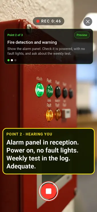 A fire alarm control panel on a reception wall with its power light on, with the Walk Cam point card and live transcription