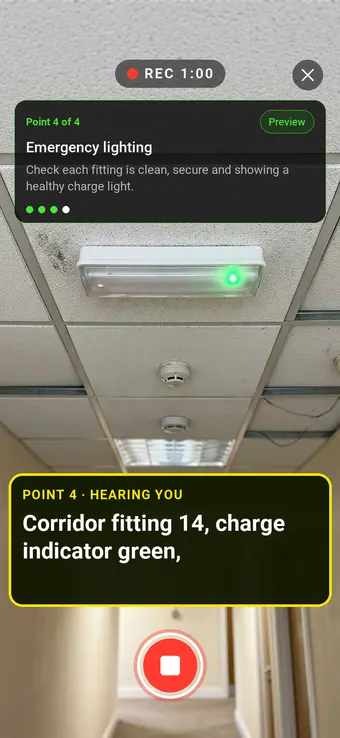 An emergency light fitting in a corridor ceiling with a green charge light, with the Walk Cam point card and live transcription