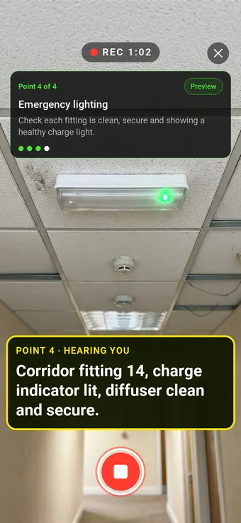 An emergency light fitting in a corridor ceiling with its charge light on, with the Walk Cam point card and live transcription
