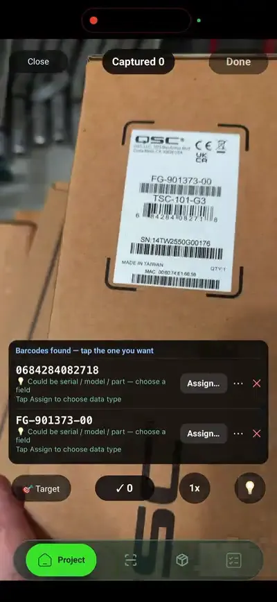 A phone held over a boxed item in a warehouse stockroom, scanning its barcode label. SiteSurvey has read the serial and part numbers off the label and is offering to assign each code to a data point on the asset record